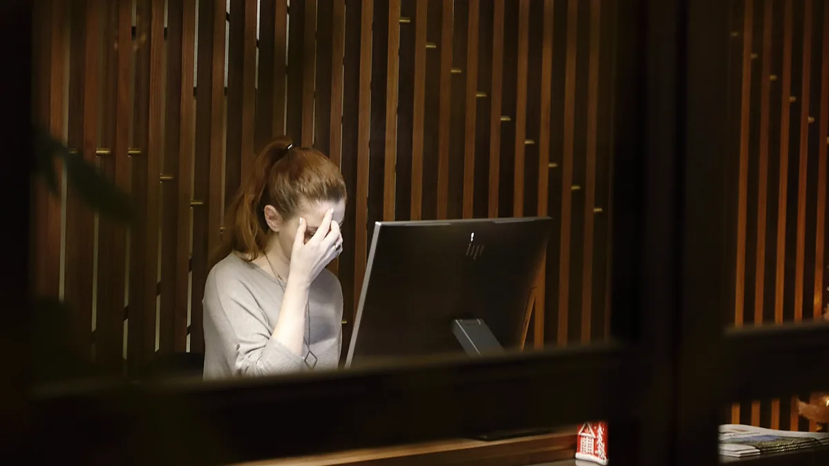 Woman at a desk behind wooden slats, head in hand